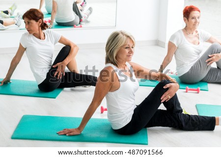 Group of middle aged women warming up in gym.Threesome sitting on floor on rubber mattresses.