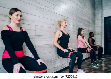 Group Of Modern Diverse Young Women Leaning On Wall In Squat Exercising With Bright Red Fitness Elastic Band Modern Studio