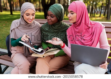 Group Of Modern Muslim Students Ladies Learning Reading Books Sitting On Bench Outside. Islamic Females Learn Together Using Laptop Discussing Something In University Campus Park