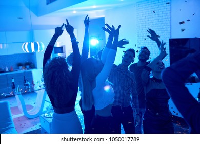 Group of modern young people dancing under confetti at private house party lit by blue light