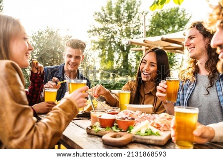 Group of multi ethnic friends having backyard dinner party together -  Diverse young people sitting at bar table toasting beer glasses in brewery pub garden - Happy hour, lunch break and youth concept