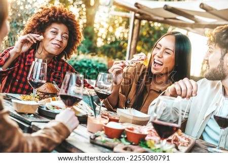 Group of multi ethnic friends having dinner party in pub garden - Happy young people drinking wine and eating food sitting in bar restaurant table - Friendship, lunch break and youth culture concept