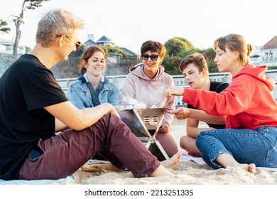 Group Of Multiaged Friends And Family Toasting Marshmallows Around Fire On The Beach. Active Teenagers With Parents Outdoor Activity. Grilled Sweets On The Fire. Sunset Time. Selective Focus