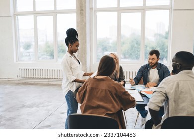Group of multiethnic colleagues gathering around table while black woman standing near table and sharing information while having discussion about project in workspace - Shutterstock ID 2337104463