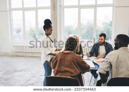 Group of multiethnic colleagues gathering around table while black woman standing near table and sharing information while having discussion about project in workspace