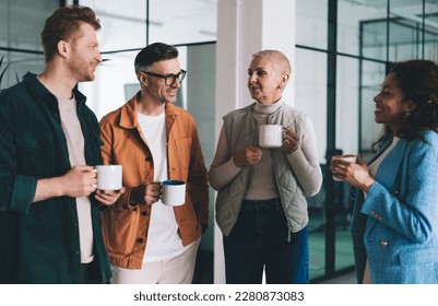 Group of multiethnic colleagues standing with cups of coffee and looking at each other while taking coffee break and speaking about business strategy steps going forward in modern office - Powered by Shutterstock