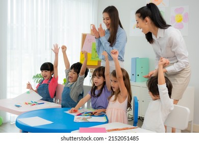 A Group Of Multi-ethnic Cute Preschool Children, Boys And Girls, Sitting Drawing Happily At A Table, Raising Hands And Singing Along With A Beautiful Asian And Caucasian Teachers At A Kindergarten.