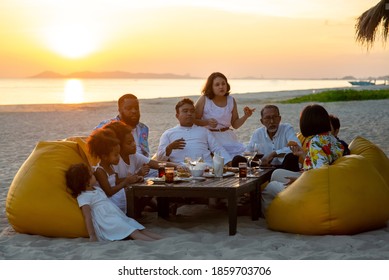 Group Of Multiethnic Family Friends Enjoy Dinner Party Together On The Beach At Sunset. Diverse Family With Child Girl, Adult And Senior Couple Relax And Having Fun Together On Summer Holiday Vacation