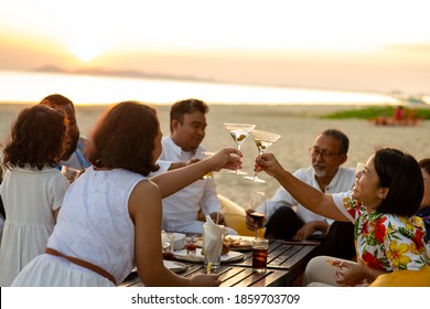 Group Of Multiethnic Family Friends Enjoy Dinner Party Together On The Beach At Sunset. Diverse Family With Child Girl, Adult And Senior Couple Relax And Having Fun Together On Summer Holiday Vacation