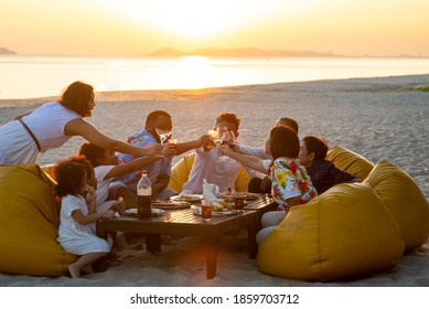 Group Of Multiethnic Family Friends Enjoy Dinner Party Together On The Beach At Sunset. Diverse Family With Child Girl, Adult And Senior Couple Relax And Having Fun Together On Summer Holiday Vacation