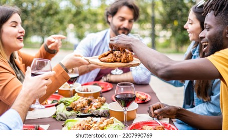 Group multi-ethnic friends having lunch at farmhouse table - Diverse young people sitting at home during bbq party -  African guy taking the grilled chicken wings by hands - Powered by Shutterstock