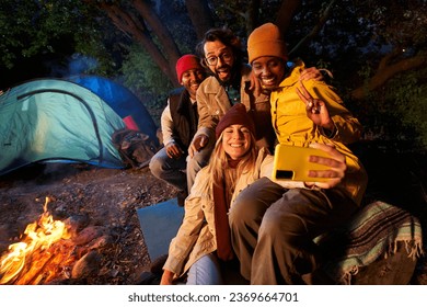 Group multi-ethnic friends smiling and taking selfie phone sitting front campfire. Young gathered together camping on winter night. Two cheerful millennial excited couples happy embracing on getaway.  - Powered by Shutterstock