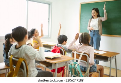Group of Multi-ethnic happy elementary school children raising their arms to answer asian teacher question in classroom. Education, elementary school, learning, Back to school concept