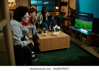 Group of multi-ethnic male friends sitting in living room watching soccer match on TV in evening, copy space - Powered by Shutterstock