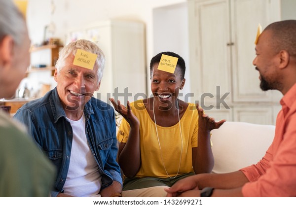 Group of multiethnic mature friends playing\
guess sticky head game at home. Men and smiling women enjoying\
charades game session. African woman trying to make her friend\
guess the character.