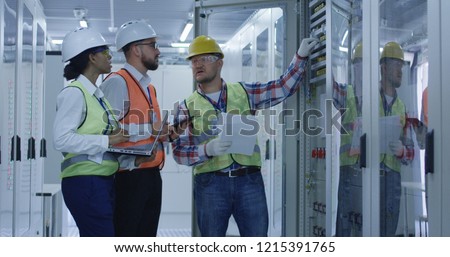 Group of multiethnic men and woman in hardhats working in hall of solar plant control center having discussion between racks
