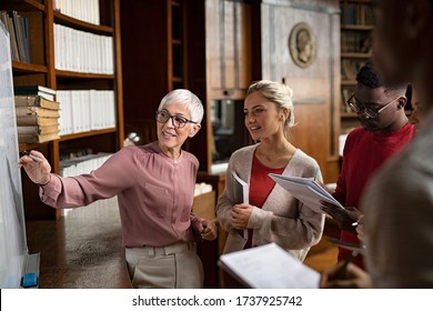 Group Of Multiethnic Students Taking An Active Part In A Lesson While Standing In Library. Mature Woman Teacher And University Student Discussing Over Math Formulas On Whiteboard. Professor Teaching.