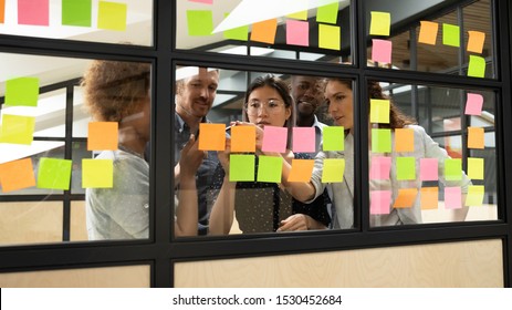 Group of multi-ethnic workers creating and assigning projects between themselves writing notes tasks using multicolored post-it sticky notes attached on glass wall concept of organization and teamwork