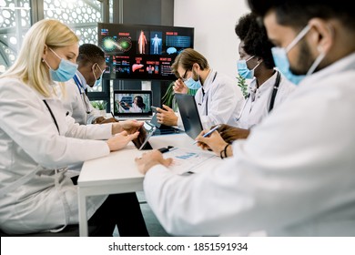 Group Of Multiethnical Healthcare Workers In Masks, With Digital Tablets And Laptop, Meeting In Hospital Boardroom. Medical Staff During Morning Online Briefing With Their Chief Doctor, African Lady