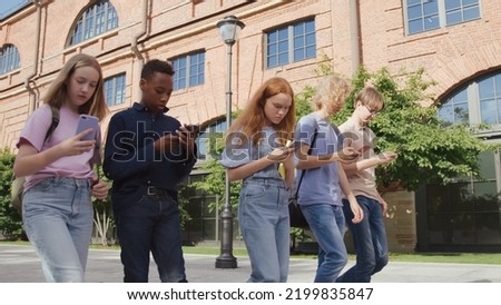 Group of multinational college students obsessed with smartphones walking outdoors. Multiethnic teens use cellphone walking together after school