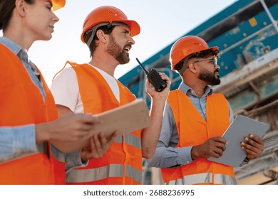 Group of multinational construction engineers in protective gear analyzing project at worksite using walkie talkie wearing protective helmet and orange vest. Teamwork concept