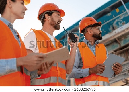 Group of multinational construction engineers in protective gear analyzing project at worksite using walkie talkie wearing protective helmet and orange vest. Teamwork concept