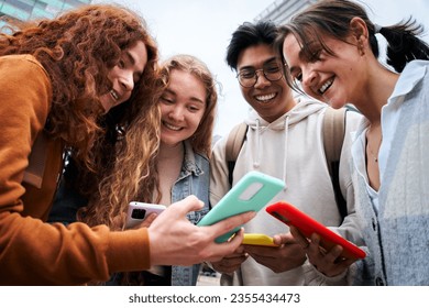 Group of multiracial classmate students looking at their smartphones sharing social media. Cheerful gathering outside university during daytime. Friends generation z having fun using mobile phone