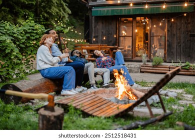 A group of multiracial friends relax around a campfire, laugh and enjoy the cozy atmosphere of a rustic outdoor setting, illuminated by string lights. - Powered by Shutterstock