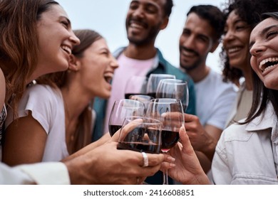 Group multiracial laughing friends toasting glasses red wine and celebrating party outdoors. Young people together cheers on open air. Boys and girls enjoying free time on summer weekend vacation.