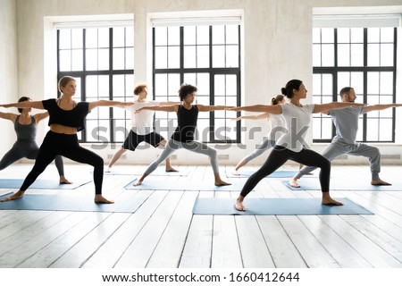 Group of multiracial people practicing yoga asanas, doing Warrior Two Virabhadrasana 2, work out indoors full length, posture increases stamina and flexibility, improves physical and mental endurance