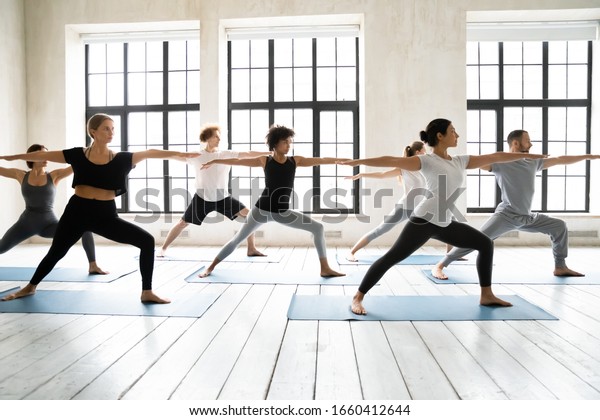 Group of multiracial people practicing yoga\
asanas, doing Warrior Two Virabhadrasana 2, work out indoors full\
length, posture increases stamina and flexibility, improves\
physical and mental\
endurance
