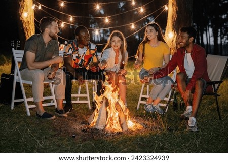 Group of multiracial restful friends roasting marshmallows while sitting by bonfire
