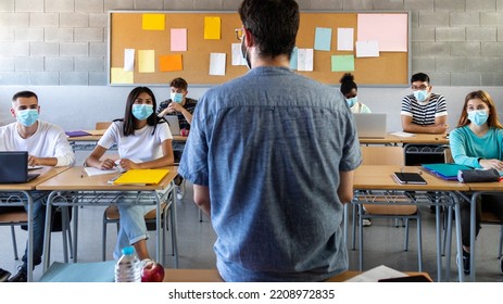 Group Of Multiracial Teen High School Students Wearing Face Mask Listen To Teacher Lesson. Rear View Of Teacher.