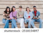 Group of multiracial young friends sitting on a staircase using their cellphones to share photos on social media. Teenage student people playing a game together with a mobile phone app on a meeting