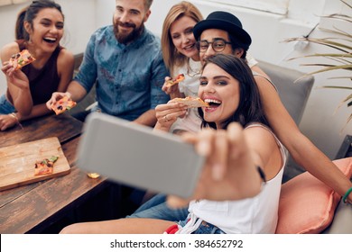 Group of multiracial young people taking a selfie while eating pizza. Young woman eating pizza her friends sitting around during a party. - Powered by Shutterstock