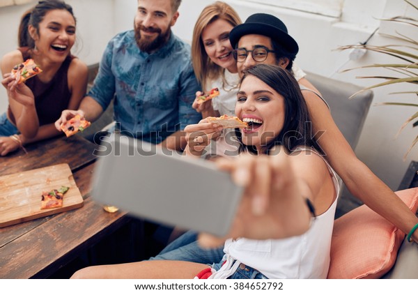 Group of multiracial young people taking a selfie\
while eating pizza. Young woman eating pizza her friends sitting\
around during a party.