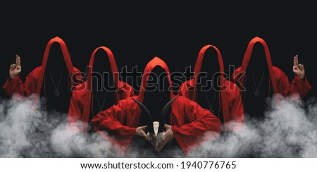 Group of mysterious figures in hooded cloaks in the dark. Leader of sectarians holds skull with horns. Horror scene with smoke. Black background.