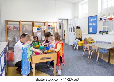 Group Of Nursery Children In A Classroom. Most Are Playing At The Water Table And Three Children Are Playing In The Sand Table.