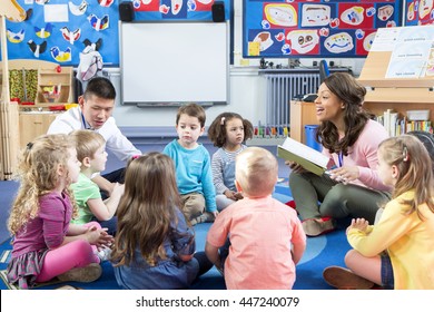 Group Of Nursery Children Sitting On The Floor In Their Classroom With Their Teachers. The Female Teacher Is Reading From A Book. 