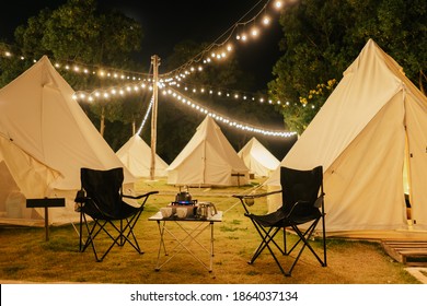 Group outdoor camping teepee tent and night light with two empty chairs with picnic table and accessories in the forest. Glamping camping tent in the forest under night sky.