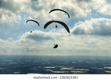 A group of paragliders soars through a dramatic cloudy sky, illuminated by breaking sun rays. The silhouetted figures fly high above a vast expanse of agricultural fields and rural plains. This sweeping aerial view captures the thrill of extreme outdoor sports, a sense of absolute freedom, and the beauty of flying over the peaceful countryside.