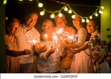 Group Of People Celebrate An Event Like New Years Eve Or Birthday All Together With Sparkles Light By Night In The Dark. Smiles And Having Fun In Friendship For Different Ages Men And Women At Home.