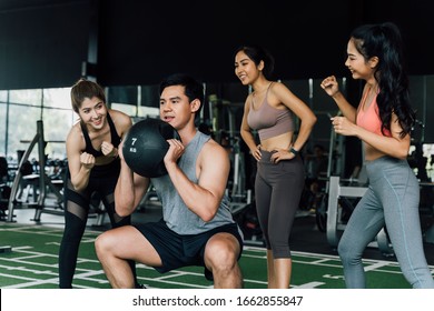 Group Of People Cheering On Their Asian Chinese Male Friend Doing Squats With A Medicine Ball In Fitness Gym. Working Out Together As A Teamwork. Encouragement And Togetherness Concept