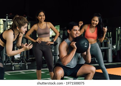 Group Of People Cheering On Their Asian Chinese Male Friend Doing Squats With A Medicine Ball In Fitness Gym. Working Out Together As A Teamwork. Encouragement And Togetherness Concept