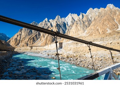 A group of people crossing a suspension bridge over a river surrounded by towering mountains at dusk, with distant peaks illuminated by the setting sun. - Powered by Shutterstock