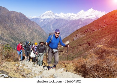 Group Of People Of Different Age And Ethnicity Walking Up On Mountain Trail During Hike In Nepalese Himalaya Carrying Heavy Backpacks And Climbing Gear Led By Mature Guide Sun Shining