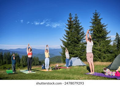 Group of people doing yoga pose outdoor in camping in the mountains. Adults and children standing on yoga mats, each doing yoga pose under clear blue sky in morning. - Powered by Shutterstock