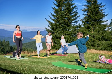 Group of people doing yoga pose outdoor in camping in the mountains. Adults and children standing on yoga mats, each doing a yoga pose under a clear blue sky in the morning. Young boy is instructor. - Powered by Shutterstock