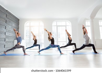 Group of people doing yoga warrior pose at white studio with gesture of will. Fitness class, sport and healthy lifestyle concept