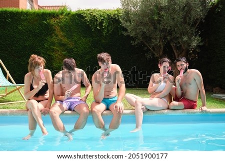 Group of people eating strawberry popsicles sitting near by a swimming pool.